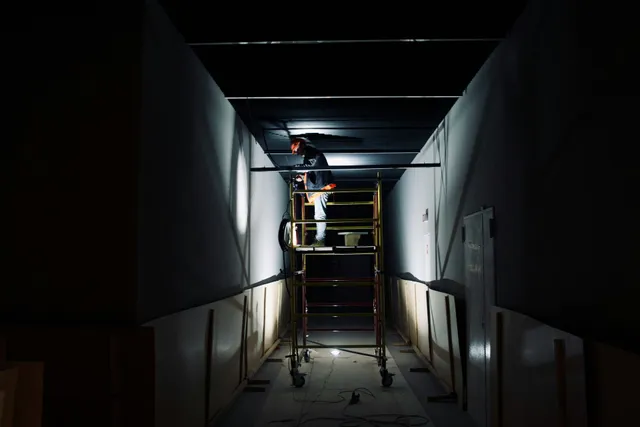 Worker on a scaffold in a dim hallway, installing ceiling panels with a flashlight.