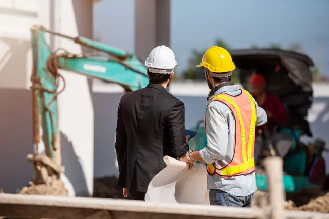 Two construction workers in hard hats reviewing work near an excavator at a building site
