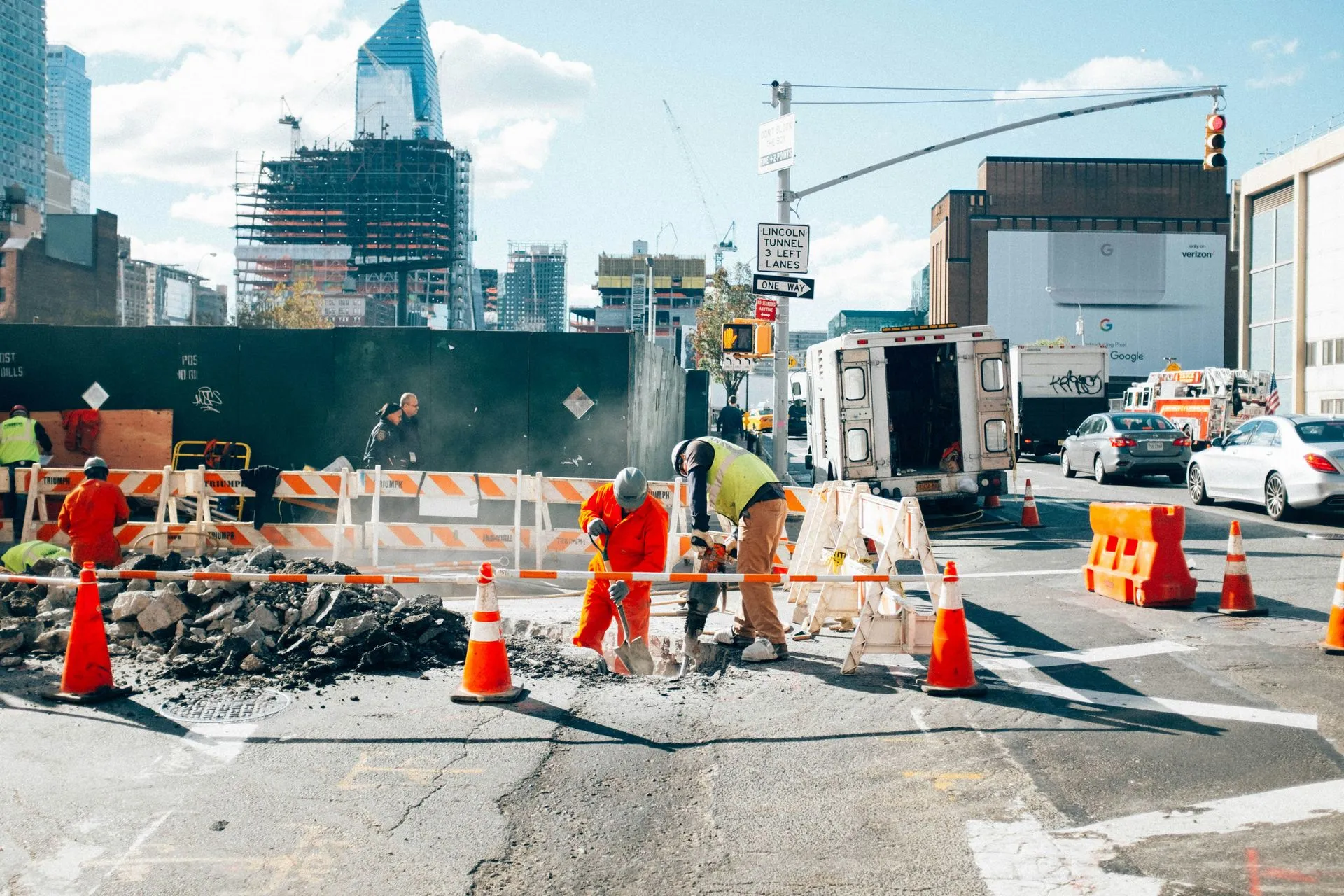 Road construction crew repairing a city street with orange cones, barriers, and traffic in the background