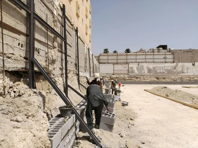 Two people walk along a gravel path beside a high stone wall and metal railings in a sunlit ruin.