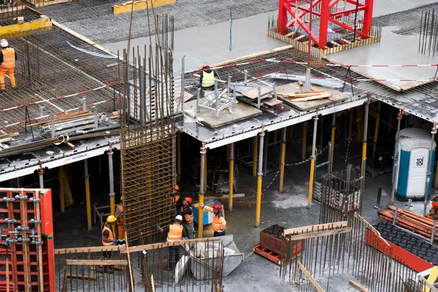 Construction workers on a rebar and concrete job site beneath scaffolding and yellow supports
