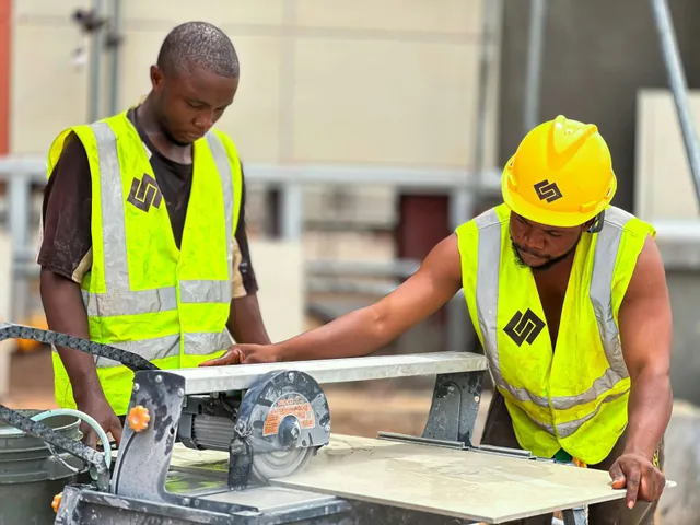Two construction workers in yellow safety vests and hard hats measuring wood with a circular saw on a worksite