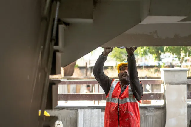 Construction worker in a red vest and yellow hard hat lifting an overhead panel outdoors