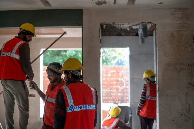 Construction workers in red vests and yellow hard hats inside an unfinished building, working on walls.