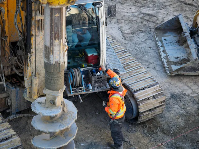 Construction workers inspect a drilling rig beside a large auger on a muddy site.