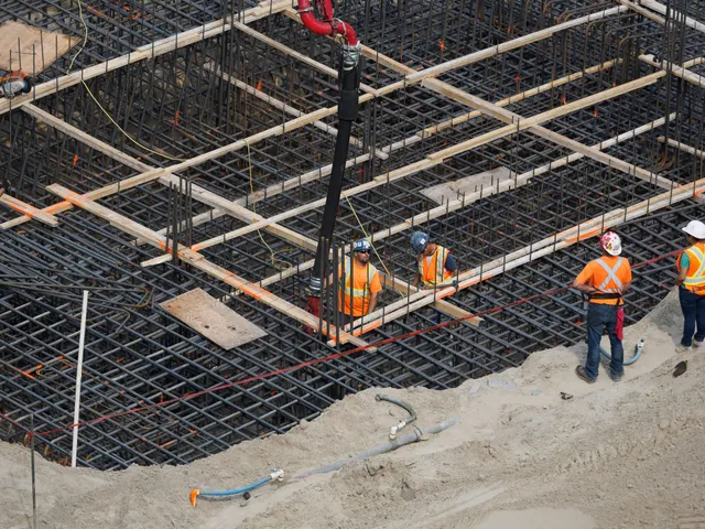 Construction workers in orange vests on rebar foundation with concrete pump hose