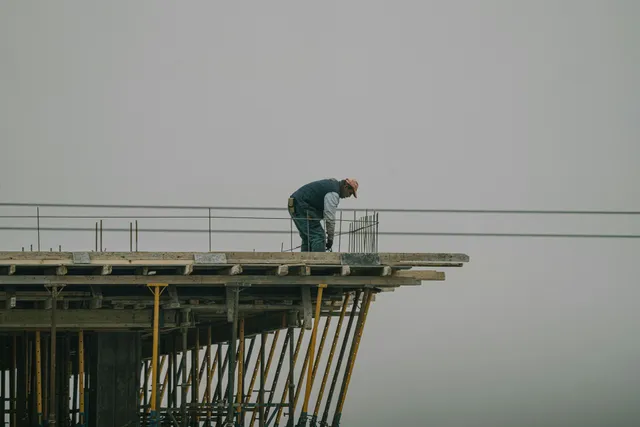 Worker bent over on a construction platform high above the ground, with scaffolding against a gray sky
