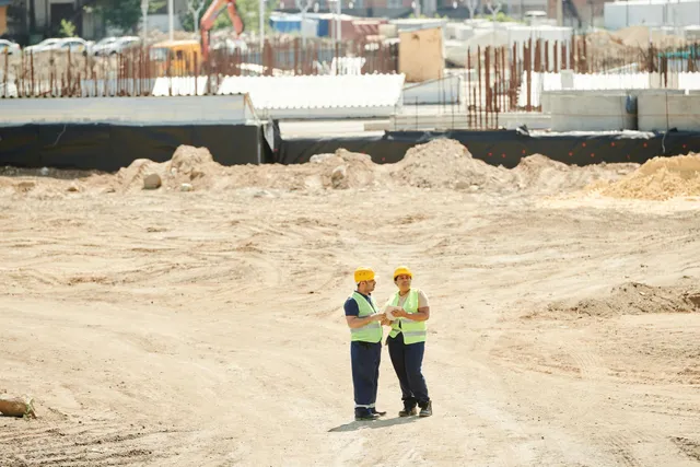 Two construction workers in hard hats stand on a sandy construction site with heavy machinery in the background