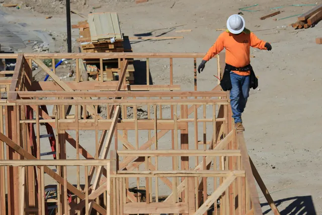 Construction worker in orange shirt and white hard hat walking beside wooden building frames on a dusty site