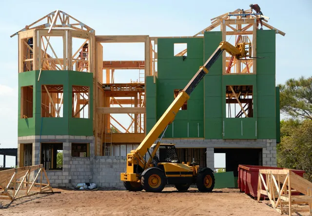 House under construction with green exterior panels and a yellow crane lifting materials.