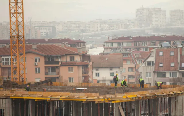 Construction workers on a high-rise slab with a crane, overlooking apartment buildings in hazy cityscape
