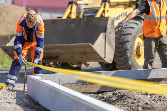 Construction workers pouring concrete into a curb form beside a yellow loader at a work site