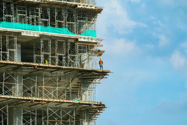 Construction site with workers on scaffolding beside a partially built high-rise against a blue sky