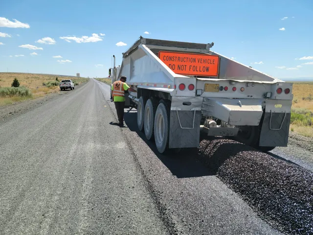 Road worker beside dump truck spreading gravel on a rural dirt road under a clear blue sky