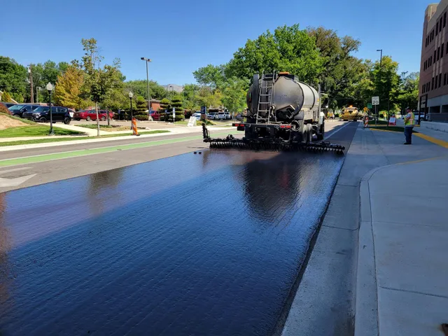 Road paving machine laying fresh black asphalt on a sunny street beside a sidewalk and trees