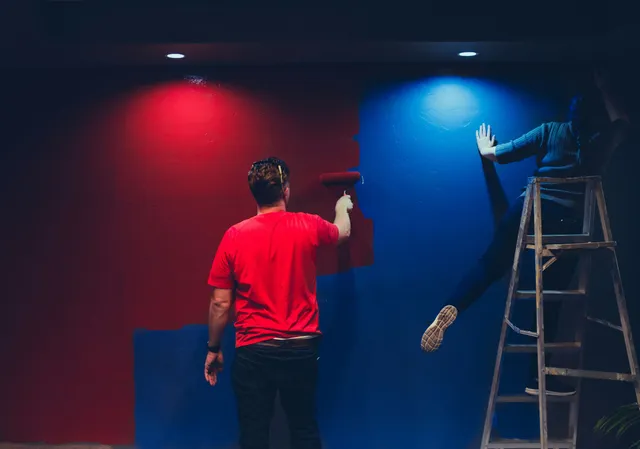 Person in red painting a wall under red and blue lights; another person stands on a ladder.