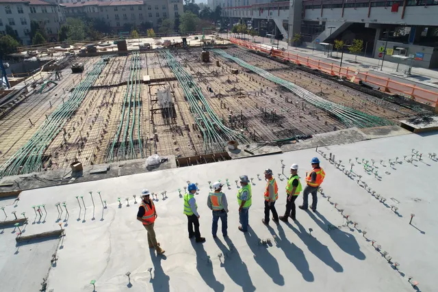 Construction workers in safety vests stand on a rooftop overlooking a large worksite.