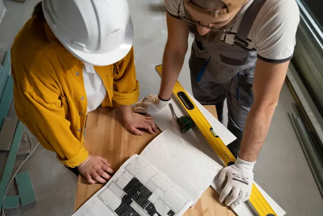 Two construction workers reviewing blueprints on a table, wearing hard hats and gloves.